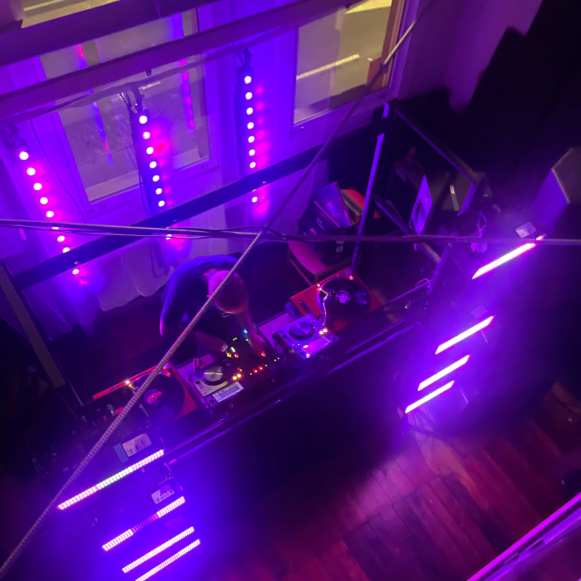 A birds-eye-view of a DJ behind a set of turntables. The room is dimly lit but they are surrounded by purple lighting strips.